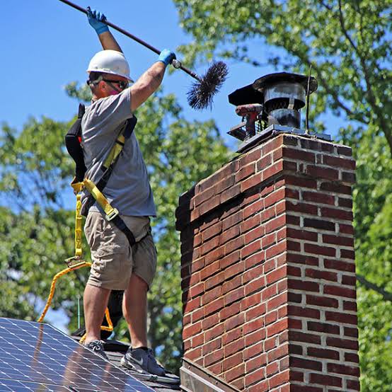 A person on a roof holding a chimney brush above a brick chimney
